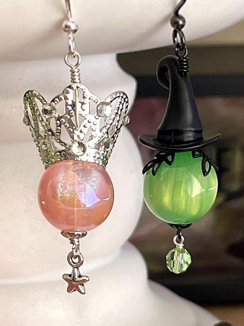 May include: A pair of whimsical earrings. One features a silver crown over a pink iridescent orb with a star charm. The other has a black witch hat atop a green orb with a green crystal charm. Both have silver hooks.