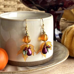 May include: Gold-toned dangle earrings with autumn-themed charms. Each earring features a pumpkin-shaped bead, a maple leaf, and a cluster of purple and red beads. The earrings are displayed near a ceramic mug and a small pumpkin.