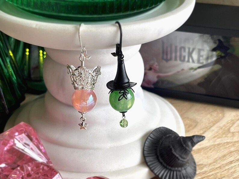 May include: A pair of whimsical earrings. One earring features a silver crown with a pink orb and star charm. The other earring has a black witch hat atop a green orb with a leaf design and a green bead charm. The word "WICKED" is visible in the background.