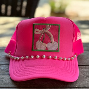 Puede incluir: Una gorra de camionero rosa con un diseño de cereza bordado en blanco y verde. La gorra tiene un borde de perlas alrededor del borde.