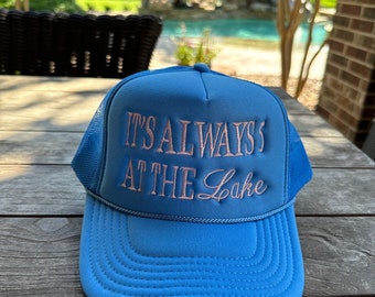 May include: A blue and black trucker hat with a pink embroidered design that says "It's Always at the Lake".