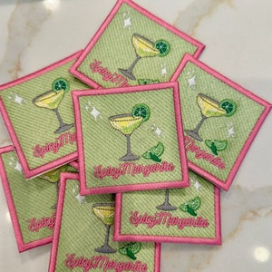 Op de afbeelding: Een stapel vierkante patches in groen en roze met geborduurde afbeeldingen van een margarita-glas en de tekst "Spicy Margarita".