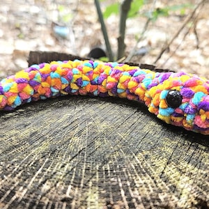 May include: A colorful, plush crochet snake toy in shades of purple, yellow, orange, and blue. The snake has a black eye and is resting on a weathered wooden stump. The background is blurred with natural foliage.