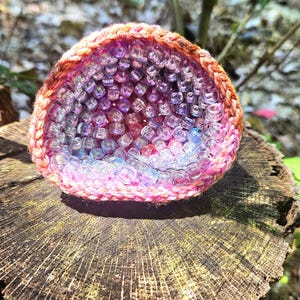 May include: A crocheted, geode-shaped object filled with clear, iridescent beads. The crochet work transitions from pink to orange, while the beads shift from pink to blue. The geode sits on a weathered tree stump, with a blurred natural background.