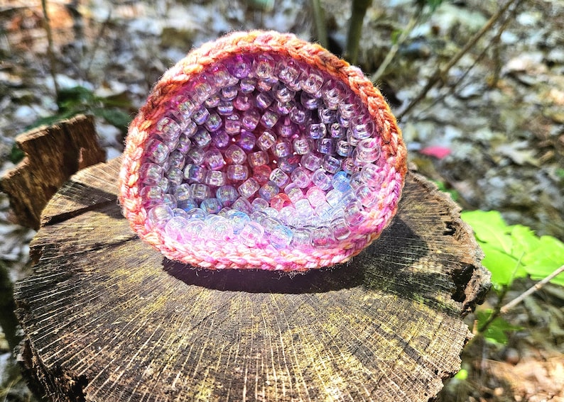 May include: A crocheted, geode-shaped object filled with clear, iridescent beads. The crochet work transitions from pink to orange, while the beads shift from pink to blue. The geode sits on a weathered tree stump, with a blurred natural background.