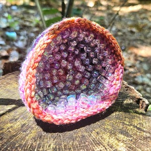 May include: A handmade, crocheted geode-shaped object filled with iridescent beads. The crochet work features a gradient of colors, including pink, orange, and purple. The geode sits on a weathered wooden surface, with natural light illuminating the piece.