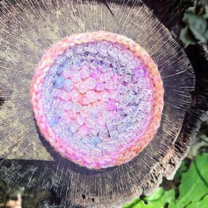 May include: A handmade, circular beaded item with a woven, pink and orange border. The interior is filled with clear, iridescent beads in shades of pink, purple, and blue. The item is set on a weathered, wooden surface.