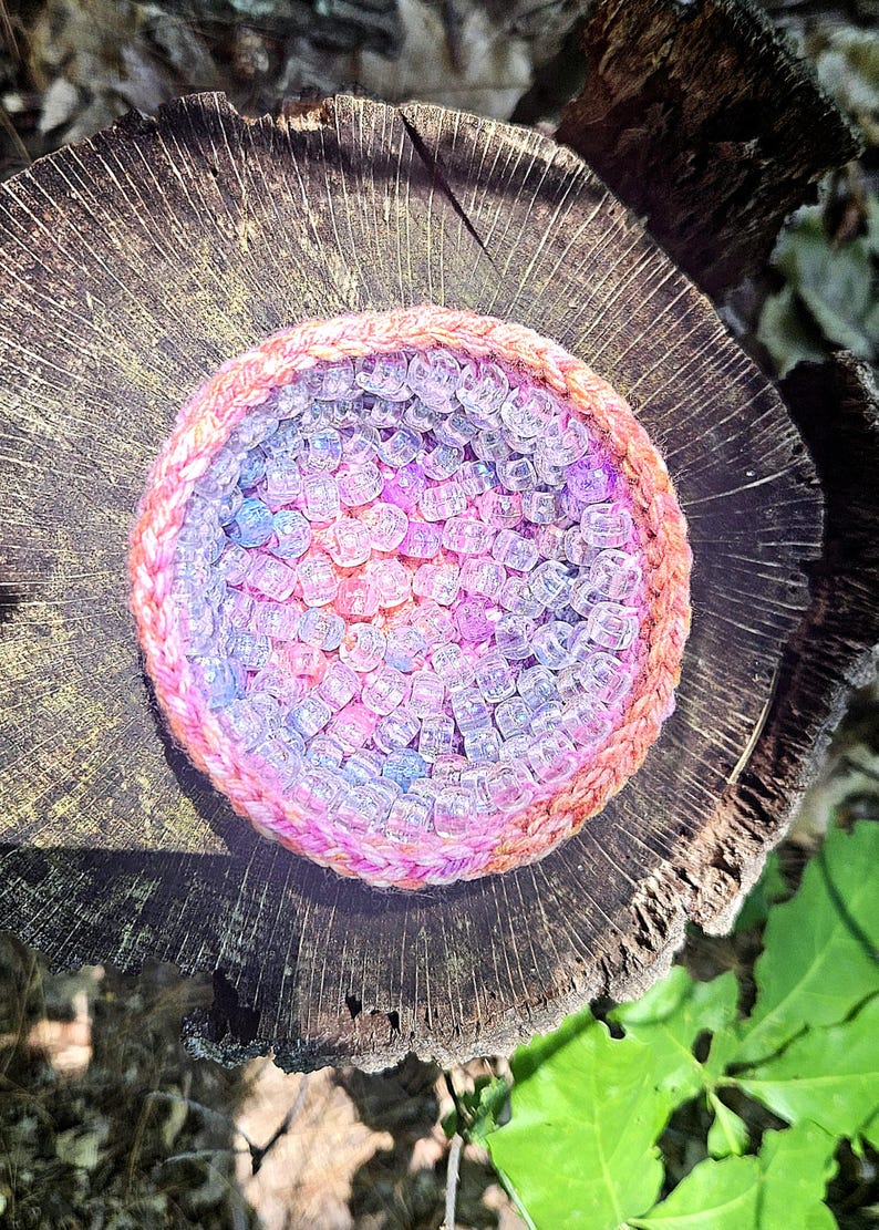 May include: A handmade, circular beaded item with a woven, pink and orange border. The interior is filled with clear, iridescent beads in shades of pink, purple, and blue. The item is set on a weathered, wooden surface.