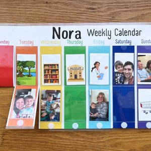 May include: A Nora Weekly Calendar with seven vertical columns in rainbow colors, each labeled with a day of the week. The calendar features clear plastic pockets containing various photo cards and illustrations, including a museum, a doctor, and a shopping cart.