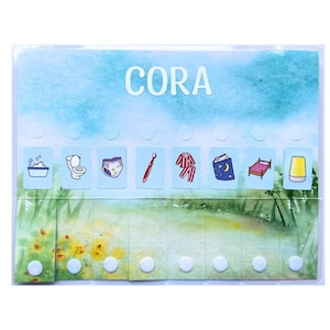 May include: A clear plastic chore chart with the name "CORA" in white letters. The chart features illustrated icons for daily tasks, including bathing, using the toilet, changing a diaper, brushing teeth, wearing pajamas, reading a book, sleeping in a bed, and drinking from a cup.
