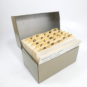May include: A gray metal index card file box with an open hinged lid. Inside are white index cards organized by beige alphabetical dividers labeled A through YYZ. The front-most card has handwritten text reading MORGIEWICZ, RALPH 102-48-4430.