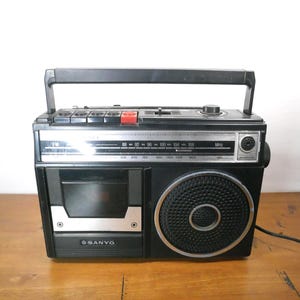 May include: A black Sanyo portable radio cassette player featuring a top carrying handle, a silver radio frequency tuning panel with FM and AM scales, a cassette deck compartment, a circular speaker grille, and various control buttons on top.