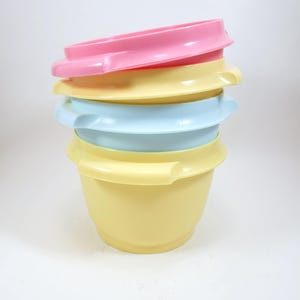 Puede incluir: Una pila de cuatro cuencos de plástico de colores con tapas. Los cuencos son de color amarillo pastel, azul y rosa. Cada cuenco tiene un asa a cada lado. Las tapas son del mismo color que el cuenco de abajo.