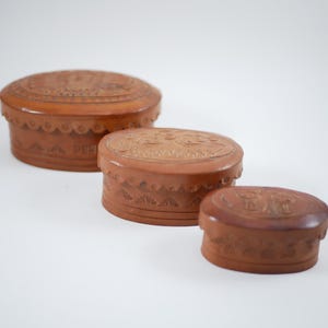 Könnte beinhalten: Drei braune, ovale Schachteln mit geschnitzten Blumendesigns auf Deckeln und Seiten. Die Schachteln sind gestapelt und werden kleiner. Die Schachteln haben einen gewellten Rand. Die Schachteln bestehen aus hellbraunem Material.