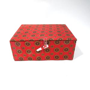May include: A rectangular red box with a repeating pattern of gold and brown circular and square designs. The box has a hinged lid and a clear plastic clasp with a white and red ribbon closure. The box is likely for storage or decorative purposes.