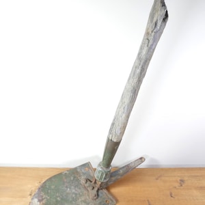 May include: A vintage folding shovel featuring a weathered, light-coloured wooden handle attached to a green, distressed metal spade head. The tool includes a metal collar and a hinged mechanism that allows the blade to be adjusted at an angle.