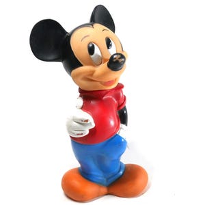 Puede incluir: Una figura vintage de Mickey Mouse, con camisa roja, pantalones azules y zapatos naranjas. El icónico personaje tiene orejas negras y una expresión sonriente. La figura es un objeto de colección.