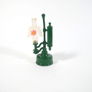 May include: A miniature green lamp with a white lampshade featuring a pink flower design. The lamp has a detailed, ornate base and a tall, slender structure. The lamp is a decorative item.