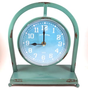 Puede incluir: Un reloj de estilo vintage en color turquesa con esfera azul claro y números blancos. El reloj indica "Antiques" y "1870". El reloj está sostenido por un soporte turquesa con una parte superior arqueada y un acabado envejecido.