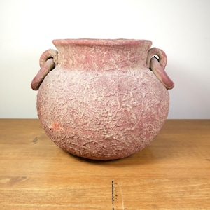 May include: A textured, round, terracotta-colored vase with two loop handles. The vase has a wide opening and a rough, aged appearance. The background is a plain white wall and a wooden surface.