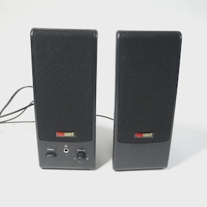 Pode incluir: Dois alto-falantes de computador Gigaware pretos. Cada alto-falante tem formato retangular com uma frente de malha preta. O alto-falante esquerdo tem um botão de energia, uma tomada para fone de ouvido e controle de volume. O logotipo Gigaware é vermelho e amarelo.