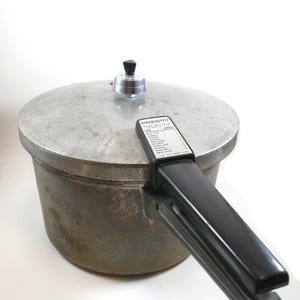 Puede incluir: Olla a presión Presto vintage con tapa plateada y asa negra. La olla tiene un aspecto desgastado y envejecido. El asa tiene una etiqueta con instrucciones de cocción. Un electrodoméstico de cocina clásico.
