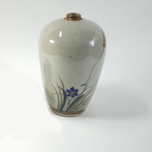 May include: A ceramic vase with a light beige glaze. The vase features a hand-painted design of a blue flower, green grass, and brown accents. The vase has a brown rim and a narrow opening.