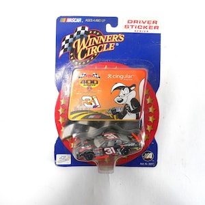 May include: A NASCAR Winner's Circle Driver Sticker Series toy car, featuring a black, red, and gray design with the number 31. The packaging includes a cartoon skunk and the text "Cingular". The package is blue with a checkered flag.