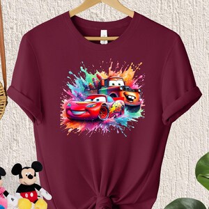 May include: Burgundy t-shirt with a colorful graphic of cartoon cars from the movie Cars. The graphic features Lightning McQueen, Mater, and a red race car.