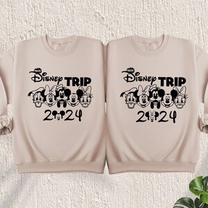 Puede incluir: Dos sudaderas color beige con personajes de Disney negros y el texto "Disney Trip 2024".