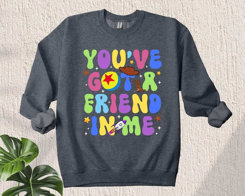 May include: A dark gray sweatshirt with the text "You've Got a Friend in Me" in colorful, retro-style letters. The text is surrounded by stars and includes a cartoon image of Woody's hat, boot, and Buzz Lightyear's rocket.