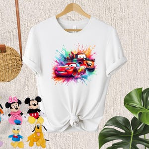 May include: White t-shirt with a colorful graphic of three cartoon cars, including Lightning McQueen, Mater, and a red race car. The graphic is surrounded by colorful paint splatters.
