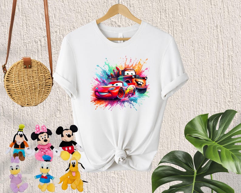 May include: White t-shirt with a colorful graphic of three cartoon cars, including Lightning McQueen, Mater, and a red race car. The graphic is surrounded by colorful paint splatters.