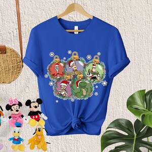 May include: Blue t-shirt with a Christmas-themed design featuring characters from the Toy Story franchise. The characters are Woody, Buzz Lightyear, Rex, and a green alien. The characters are inside of Christmas ornaments.