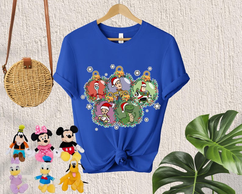 May include: Blue t-shirt with a Christmas-themed design featuring characters from the Toy Story franchise. The characters are Woody, Buzz Lightyear, Rex, and a green alien. The characters are inside of Christmas ornaments.