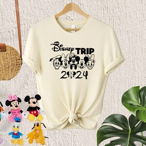 Puede incluir: Una camiseta de color crema con un gr&aacute;fico negro de Mickey Mouse y sus amigos. El gr&aacute;fico dice "Disney Trip 2024".