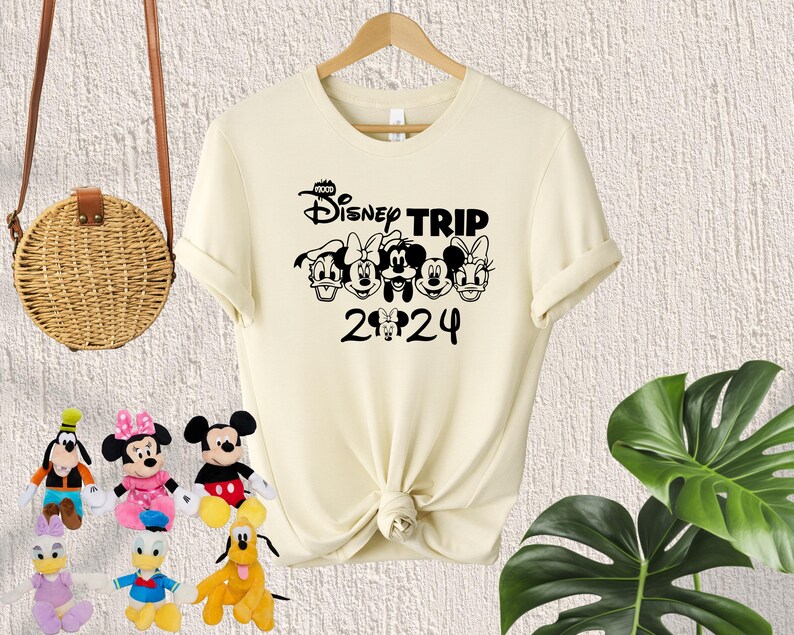 Puede incluir: Una camiseta de color crema con un gr&aacute;fico negro de Mickey Mouse y sus amigos. El gr&aacute;fico dice "Disney Trip 2024".