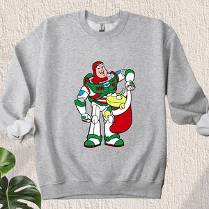 Puede incluir: Una sudadera gris con una imagen navide&ntilde;a de Buzz Lightyear de Toy Story con un gorro de Pap&aacute; Noel y sosteniendo un extraterrestre verde.
