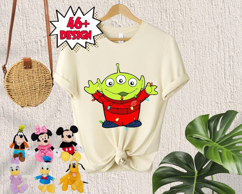 Puede incluir: Una camiseta blanca con un personaje extraterrestre verde que lleva un su&eacute;ter rojo con luces de Navidad. El extraterrestre es de la franquicia de pel&iacute;culas Toy Story. La camiseta tiene el texto "46+ Design" en un bocadillo de di&aacute;logo rojo.