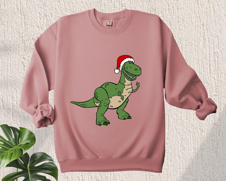 May include: A pink sweatshirt with a green dinosaur wearing a red Santa hat. The dinosaur is wearing a red Santa hat with white trim.