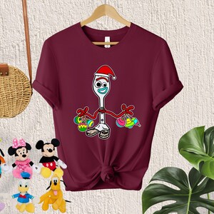 Puede incluir: Una camiseta de color granate con un contorno blanco de un personaje de juguete que lleva un gorro de Pap&aacute; Noel y sostiene adornos coloridos. El personaje es un tenedor con cara y brazos.