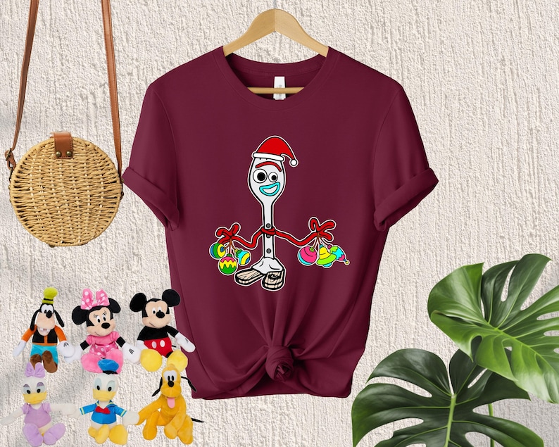 Puede incluir: Una camiseta de color granate con un contorno blanco de un personaje de juguete que lleva un gorro de Pap&aacute; Noel y sostiene adornos coloridos. El personaje es un tenedor con cara y brazos.