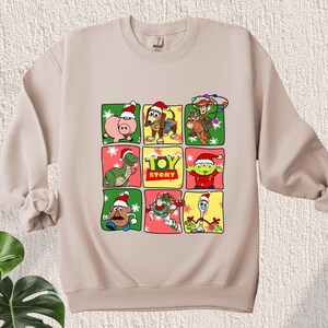 May include: Beige sweatshirt with a Christmas-themed Toy Story design. The design features characters from the movie, including Woody, Buzz Lightyear, and Rex, wearing Santa hats and surrounded by snowflakes and Christmas lights. The text "Toy Story" is printed in the center of the design.