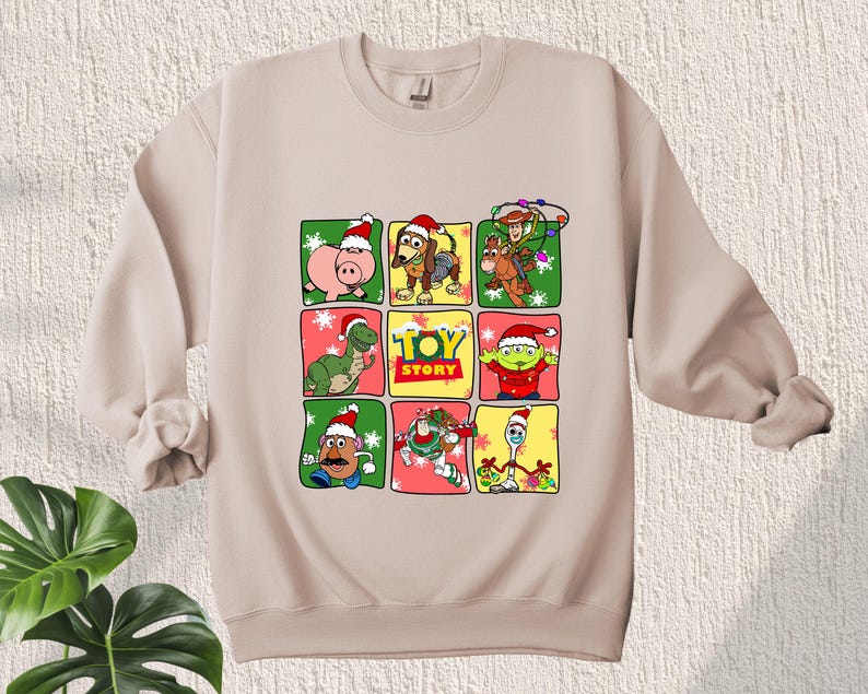 May include: Beige sweatshirt with a Christmas-themed Toy Story design. The design features characters from the movie, including Woody, Buzz Lightyear, and Rex, wearing Santa hats and surrounded by snowflakes and Christmas lights. The text "Toy Story" is printed in the center of the design.