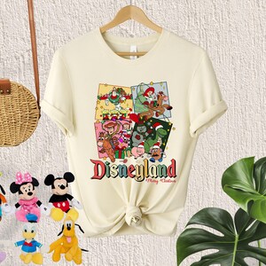 May include: A cream-colored t-shirt with a Christmas-themed design featuring characters from the Toy Story franchise. The design includes a Christmas tree, presents, and the text "Disneyland Merry Christmas".