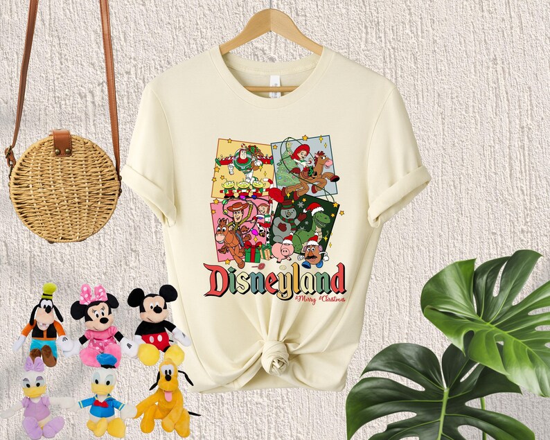 May include: A cream-colored t-shirt with a Christmas-themed design featuring characters from the Toy Story franchise. The design includes a Christmas tree, presents, and the text "Disneyland Merry Christmas".