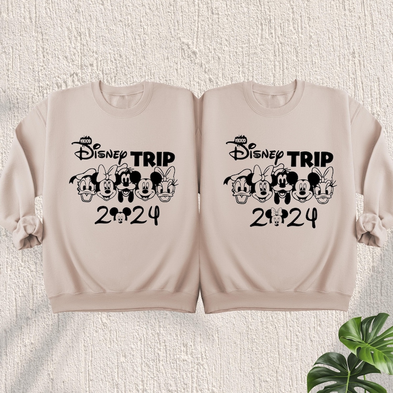 Puede incluir: Dos sudaderas color beige con personajes de Disney negros y el texto "Disney Trip 2024".