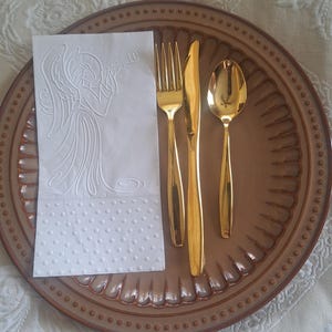 Pu&ograve; includere: Un set di forchetta, coltello e cucchiaio dorati su un piatto marrone con bordo decorativo. Un tovagliolo bianco con un disegno a rilievo di un angelo &egrave; posto sul piatto. Le posate sono disposte per una cena formale.
