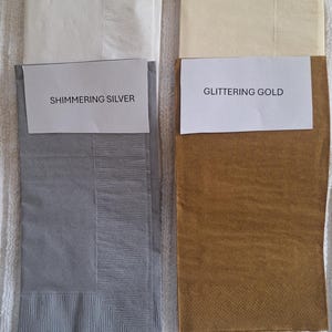 Pu&ograve; includere: Quattro tovaglioli di carta piegati in bianco, avorio, argento scintillante e oro brillante. Ogni tovagliolo ha un'etichetta con il nome del colore. I tovaglioli sono esposti su una superficie bianca strutturata.