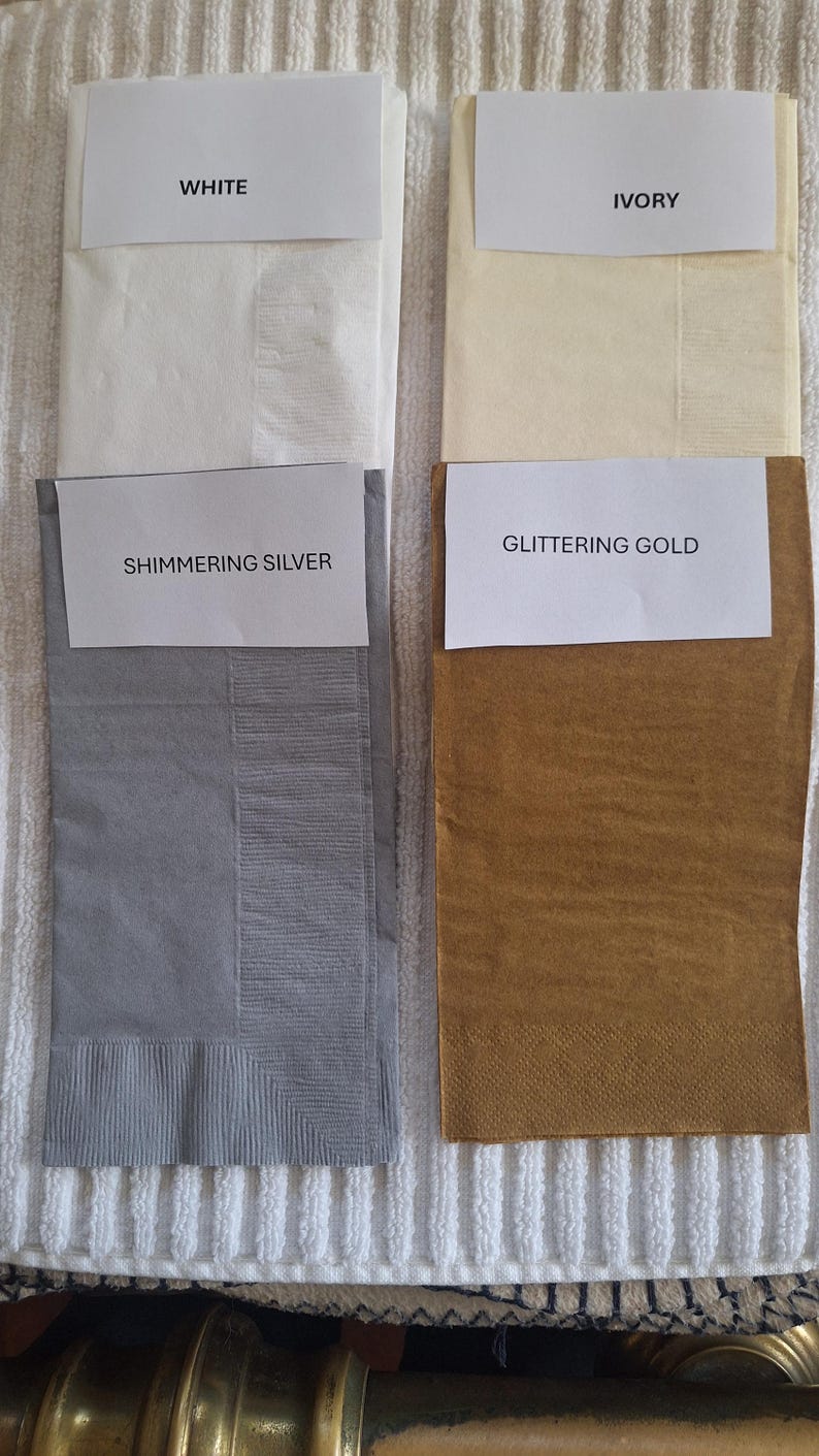 Pu&ograve; includere: Quattro tovaglioli di carta piegati in bianco, avorio, argento scintillante e oro brillante. Ogni tovagliolo ha un'etichetta con il nome del colore. I tovaglioli sono esposti su una superficie bianca strutturata.